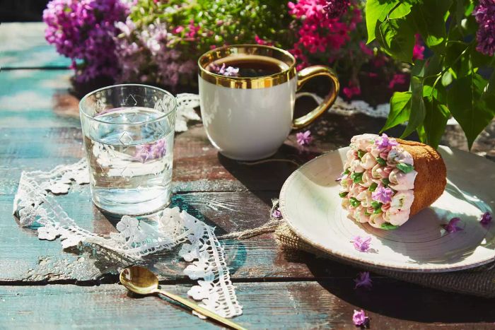A summer-themed outdoor setting featuring a coffee cup, a beautifully decorated cupcake, a glass of water, vibrant flowers, and delicate lace accents. This cottagecore-inspired picnic showcases bright floral tones under the warm glow of a sunny day, accentuated by striking shadows.