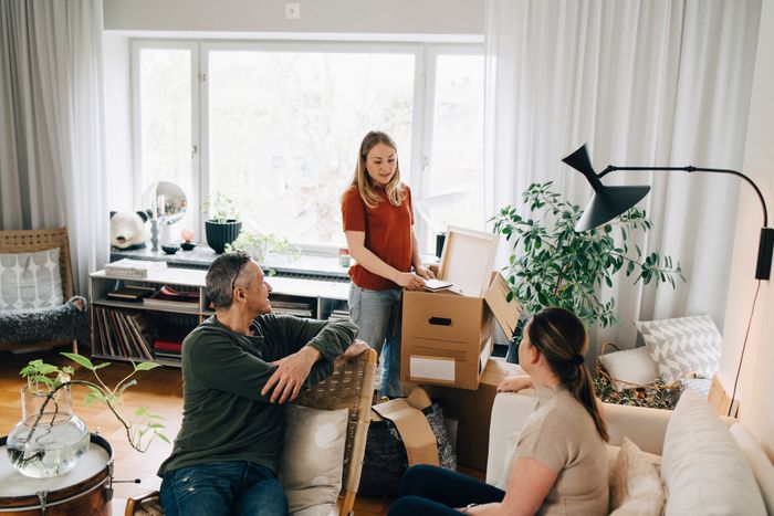 A conversation taking place in a living room surrounded by unpacked boxes, with three people appearing to plan or organize, related to work or moving topics