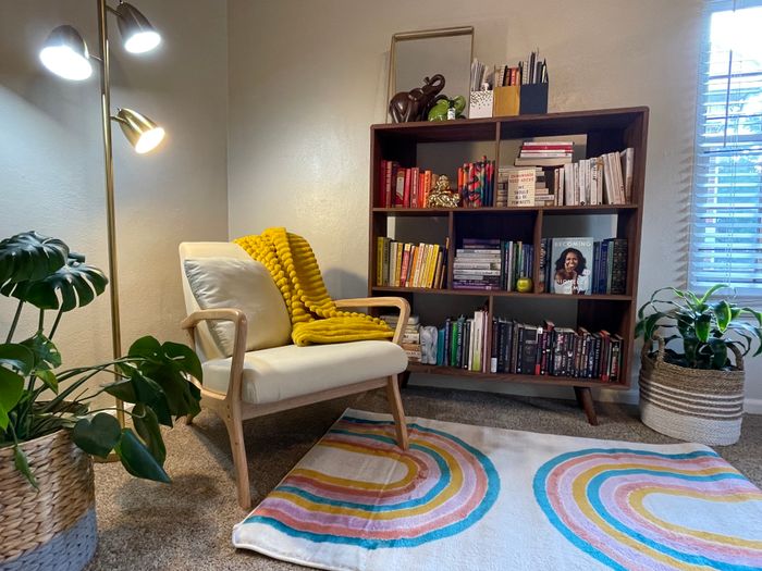 A snug reading corner featuring a plush armchair, a yellow throw blanket, a vibrant rug, bookshelves packed with books, and several potted plants
