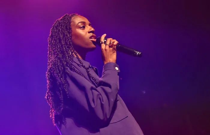 An artist stands on stage, gripping a microphone, dressed in a chic long-sleeve top, with intricately braided hair