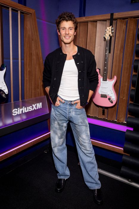 Shawn Mendes, dressed in a black cardigan layered over a white shirt and blue jeans, strikes a pose in a studio adorned with guitars on the walls
