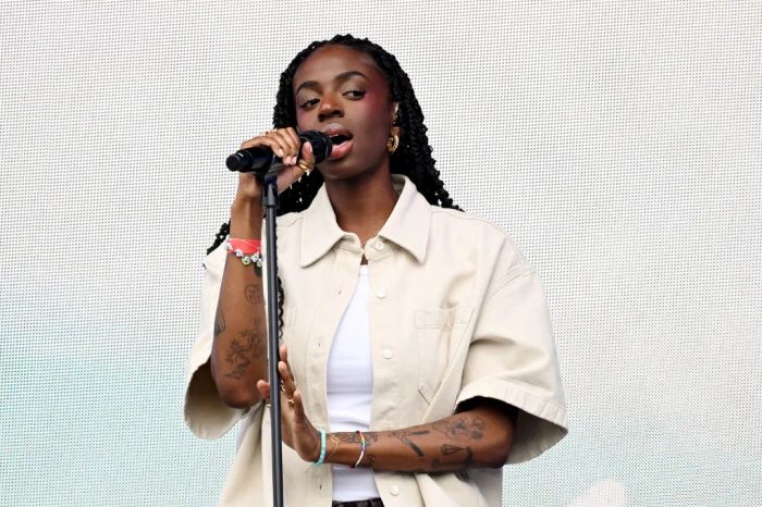 A performer with braided hair sings passionately into a microphone on stage, dressed in a short-sleeve button-up shirt, with tattoos visible on her arms