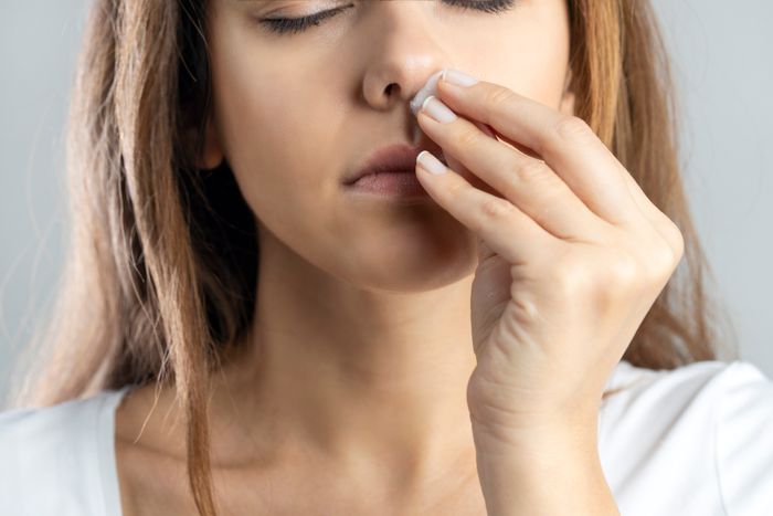 A woman delicately pinches her nose with her eyes shut, as though she is reacting to a smell or some kind of scent.