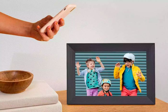 A person holding a smartphone next to the Aura Carver Smart Digital Picture Frame.