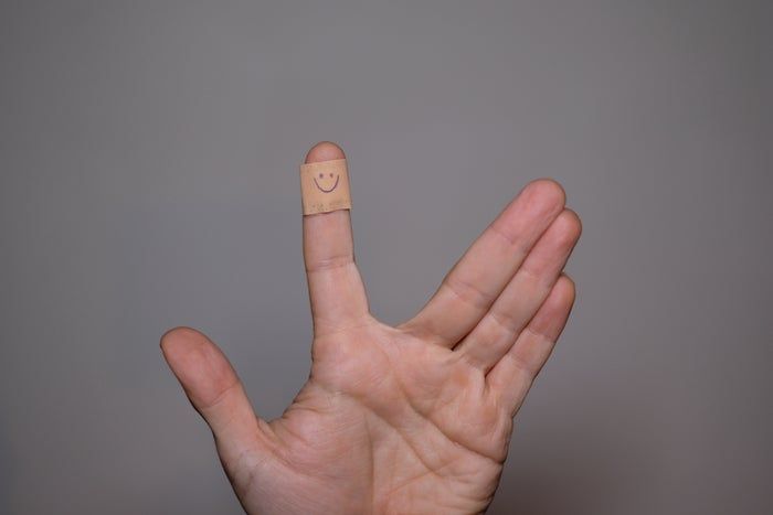 A hand wrapped with a bandage on the index finger, featuring a smiley face, showing a peace sign