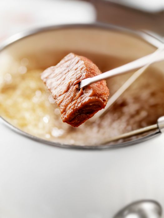 A beef cube held by tongs over a pot of bubbling hot oil, likely being cooked for fondue.