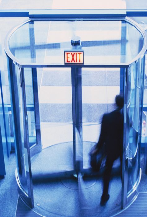 A person in a suit walking through a revolving door, viewed from behind.