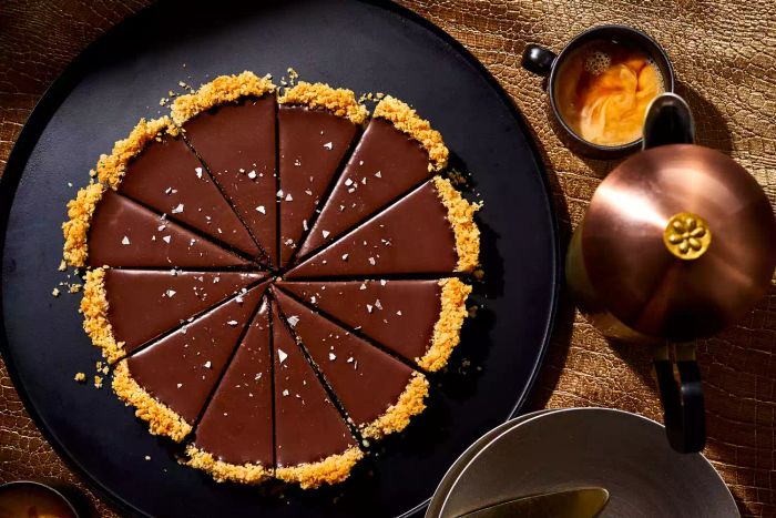 A slice of rich chocolate ganache tart rests on a sleek black plate, accompanied by a cup of coffee and a French press, creating a perfect pairing for indulgence.