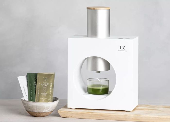 a sleek white and silver matcha machine accompanied by a cup of matcha and packets of matcha powder