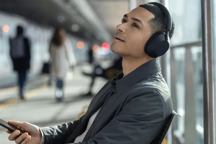 A man is sitting outdoors, wearing Sony WH-1000XM5 Noise Canceling Headphones.