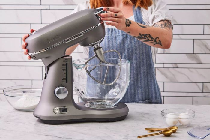 A woman is holding the top of a Breville Bakery Chef Stand Mixer placed on a white countertop.