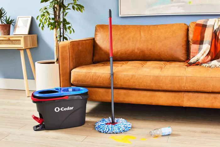 The O-Cedar EasyWring RinseClean Spin Mop & Bucket System shown on wooden flooring beside a couch and a spilled liquid.