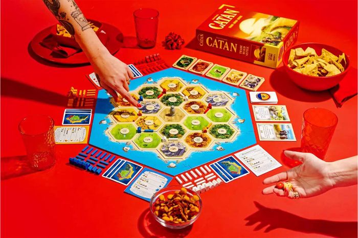 Two individuals are enjoying a round of the Catan Studio Board Game, playing with cards and snacks around them