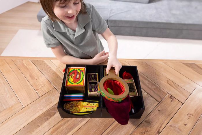 A child is holding an assortment of magic tricks, including boxes and a cloth bag, from the Melissa & Doug Deluxe Magic Set.