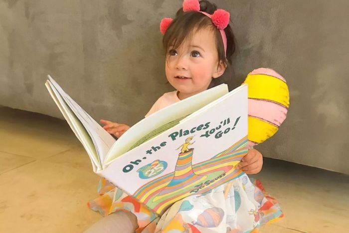 A young girl with a pink headband is holding the book 'Oh, the Places You’ll Go!' by Random House Books for Young Readers.