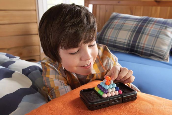 A child is engrossed in the Educational Insights Kanoodle 3D Brain Teaser Puzzle Game, lying on the bed while playing.
