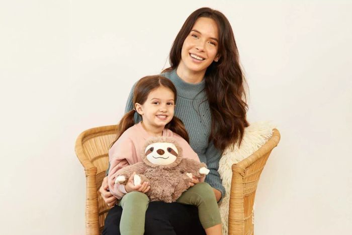 A child sits beside her mother, cuddling the Warmies Heatable and Coolable Weighted Stuffed Animal Plush