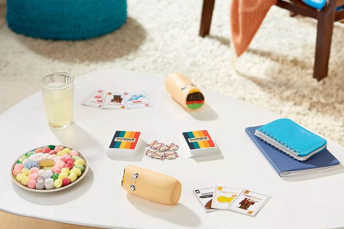 The Exploding Kittens Throw Throw Burrito Game is set up on a white table with various objects scattered around it.