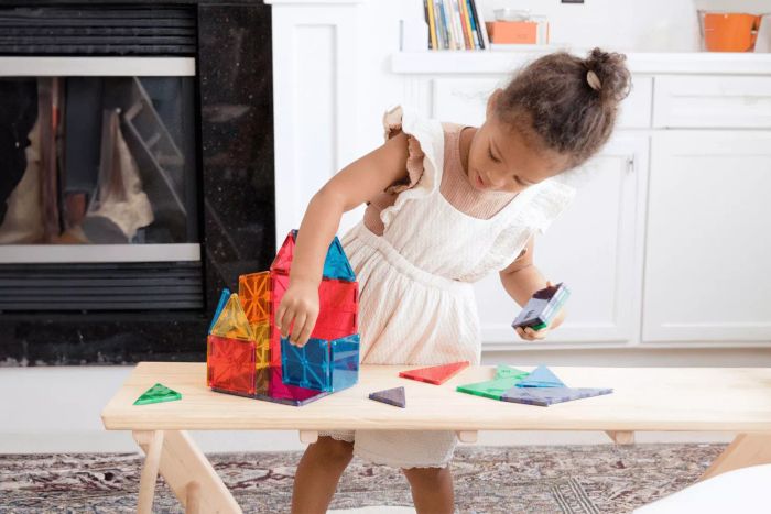 A young girl is using the Magna-Tiles 100-Piece Clear Colors Set to build a structure on a wooden table.