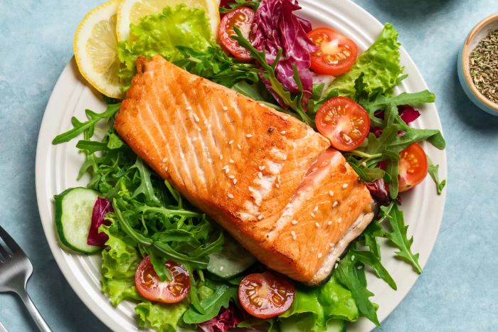 A serving of grilled salmon with a fresh vegetable salad featuring tomatoes, lettuce, and arugula. A healthy keto meal: a vibrant green salad and perfectly roasted salmon on a blue tabletop.