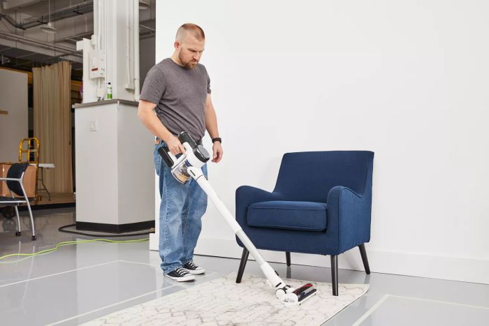 A person using the Tineco Pure One S15 Pro Cordless Stick Vacuum Cleaner to clean a rug in front of a chair.