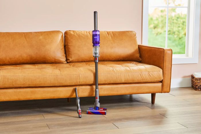 The Dyson Omni-Glide Cordless Vacuum is shown resting against a leather couch, complete with its attachment.