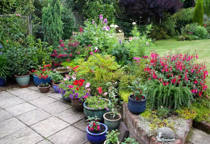 A traditional English garden with a cozy patio, vibrant flower pots, and a flowerbed teeming with colorful blossoms like fuchsias, cosmos, dahlias, geraniums, roses, hibiscus, calibrachoa, and lupins, in a horizontal format. Location: Haslemere, Surrey, England, UK.