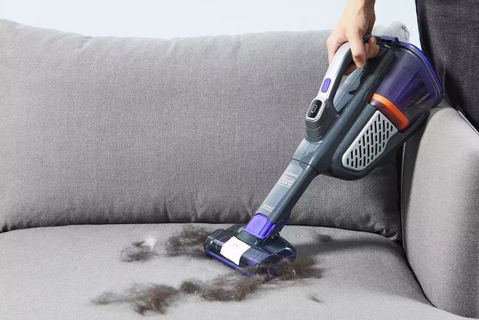 A hand is using the Black+Decker Furbuster Handheld Vacuum for Pets to remove pet hair from a gray sofa.