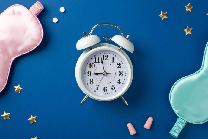 An overhead shot of a large white clock, paired with two silk sleep masks—one pink, one light blue—along with two pink earplugs, two pills, and golden star-shaped confetti scattered on a deep blue backdrop.