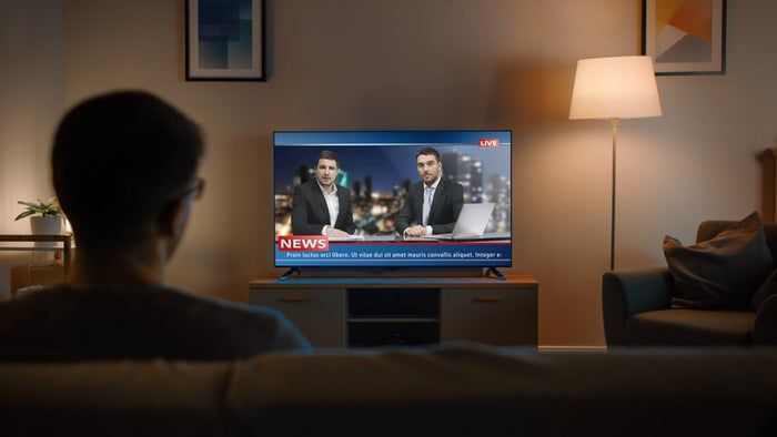 A young man wearing glasses sits on a couch, watching live news on TV.