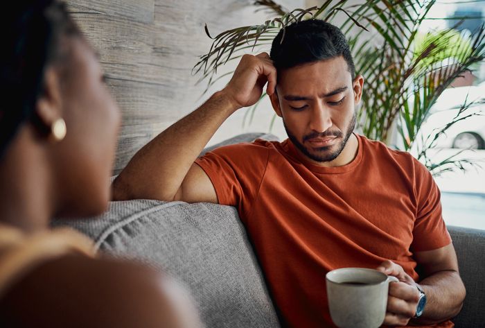 A man sitting on a couch, looking downcast, holding a cup of coffee