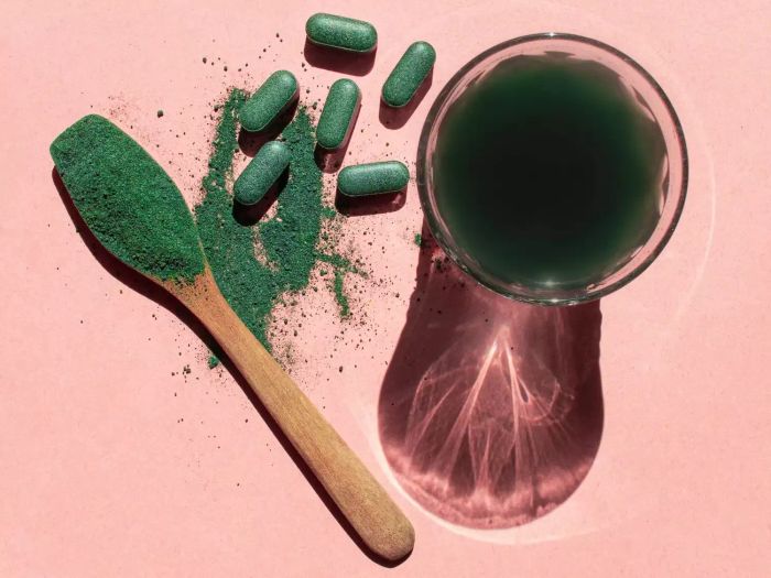 Top-down view of a glass containing chlorella drink, with spirulina and chlorella powder and tablets arranged on a pink surface.