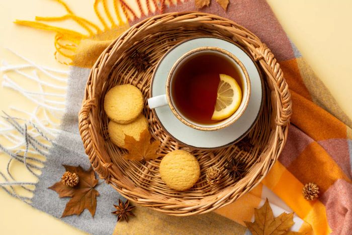 Autumn-inspired scene. A top-down shot featuring a woven tray with a cup of tea, a saucer, cookies, star anise, yellow maple leaves, pinecones, and a soft scarf on a soothing beige background.