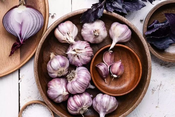 A top-down view of fresh, organic, homegrown purple vegetables and herbs, including onion, garlic, and basil, alongside cooking ingredients arranged with wooden utensils on a white wooden board. A representation of healthy eating.