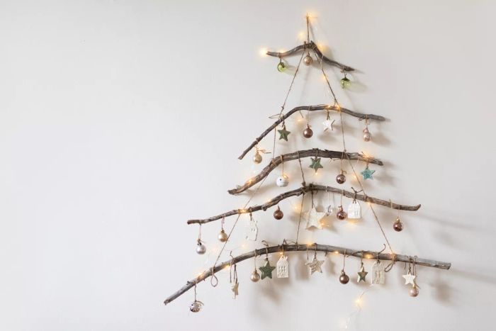 A handmade rustic Christmas tree crafted from branches, adorned with glowing garland lights and ceramic ornaments, hanging gracefully on a white wall. A DIY holiday inspiration for the Christmas and New Year season.