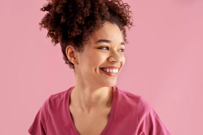 A cheerful African American woman with a genuine, radiant smile poses against a pink backdrop. A close-up shot captures a young black woman with curly hair enjoying herself in a pink studio setting, isolated for a clean background.