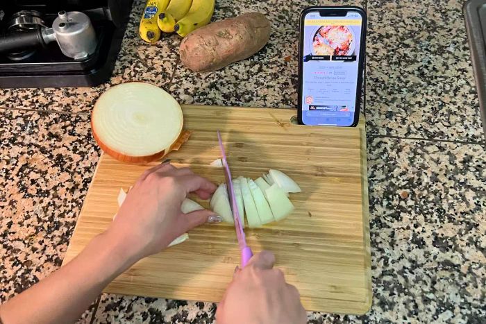 A person uses the Food52 Five Two The Happy Medium Bamboo Double-Sided Cutting Board to hold their phone while chopping an onion