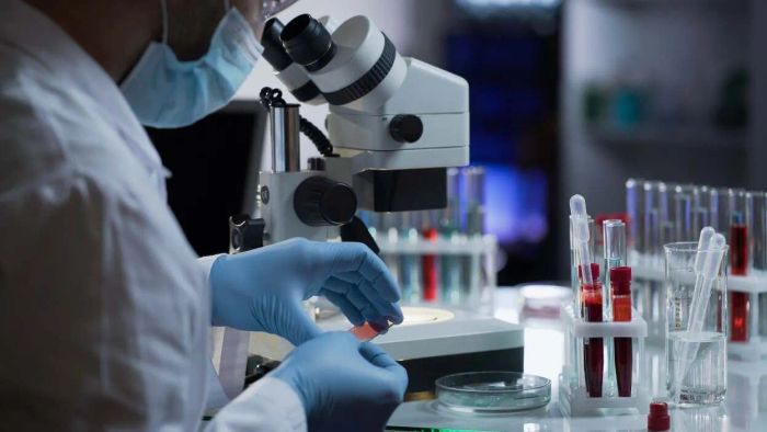 A masked laboratory technician analyzing samples using a microscope and test tubes