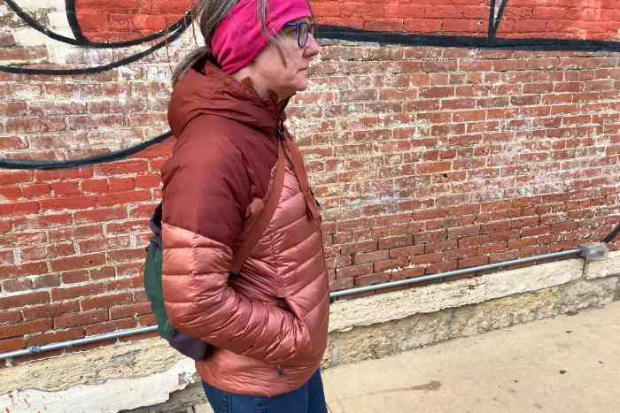 A model is seen wearing the Outdoor Research Women's Helium Down Hoodie while walking past a brick wall