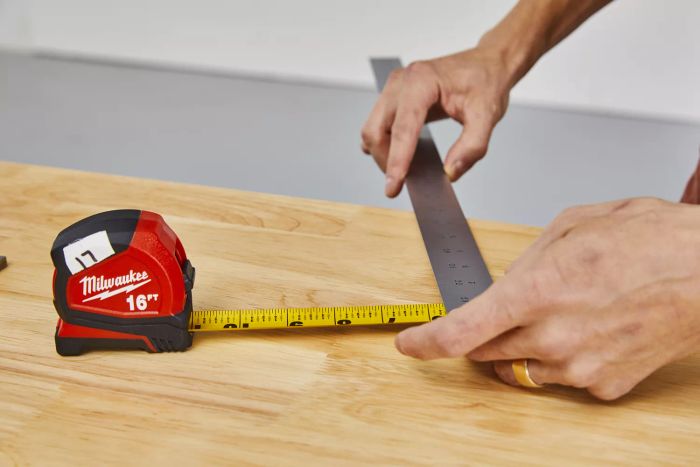 Hands measuring the width of the Milwaukee Compact Tape Measure using a metal ruler