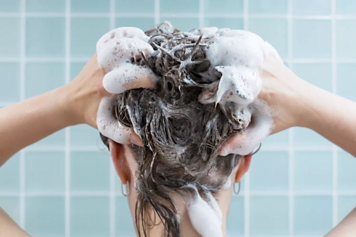 A girl is seen washing her hair with shampoo against a blue backdrop, viewed from behind
