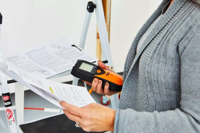 A person holding a Mecurate Stud Finder Wall Scanner along with a packet of papers