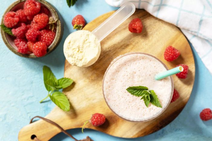 A wooden board on a stone surface displays fresh milk and a raspberry-infused protein shake with assorted berries