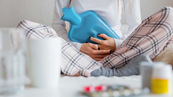 A figure in sleepwear is holding a warm water bottle, seated beside medication and glasses of water