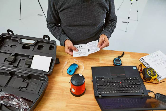 A person reviewing the instruction sheet for the Johnson Level & Tool 40-0921 Self-Leveling Cross-Line Laser Level Kit, placed next to the product case and a laptop on a wooden table