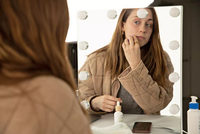 A mirror reflection of a person blending Ilia Super Serum Skin Tint With SPF 40 onto their face