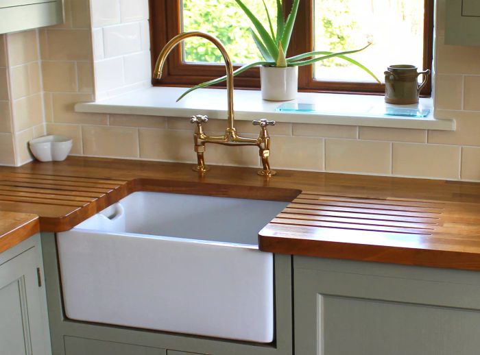 Image showing a charming farmhouse kitchen in a rustic cottage, featuring a classic white ceramic Butler / Belfast sink. The wooden cabinetry is painted in a soft green / duck egg hue, with an oak wood worktop providing a natural touch.