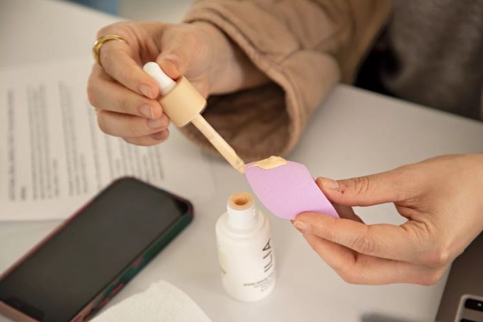 A person applying Ilia Super Serum Skin Tint With SPF 40 onto a makeup sponge beside a phone