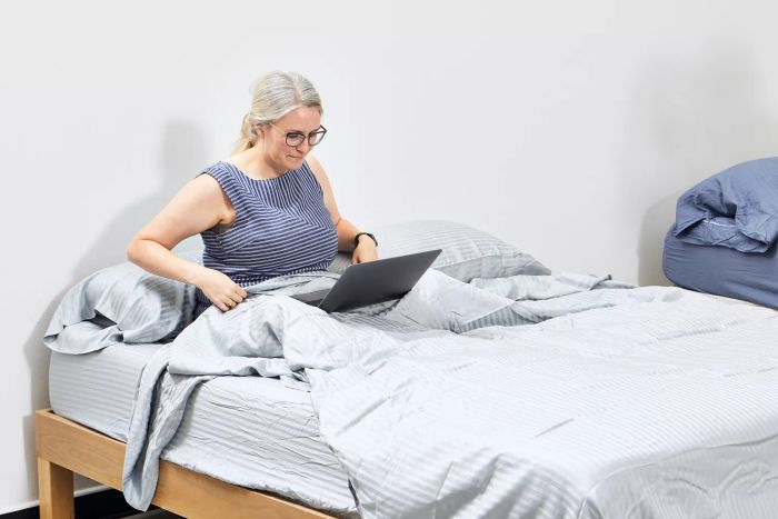 A person working on a laptop while seated on a bed dressed with the Sheets & Giggles Eucalyptus Sheet Set
