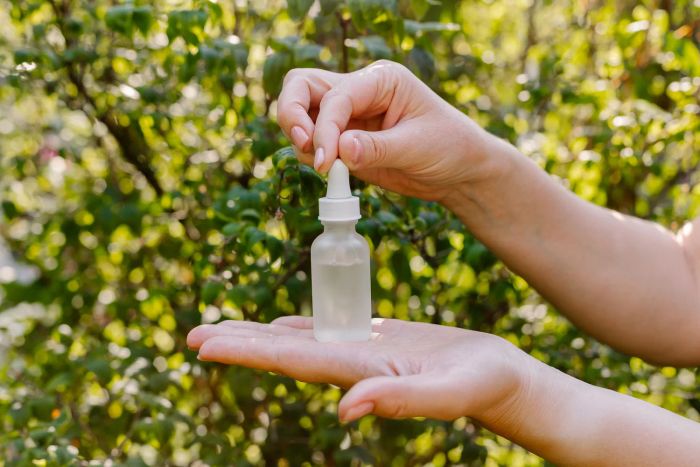 A woman holding face serum or essential oil against a lush green nature backdrop. Natural and organic skincare products for facial care.